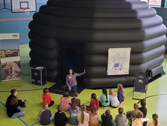 Eine Schulklasse sitzt vor dem Eingang des Pop-up-Planetariums während eine lehrende Person erklärt. (vergrößerte Bildansicht wird geöffnet)