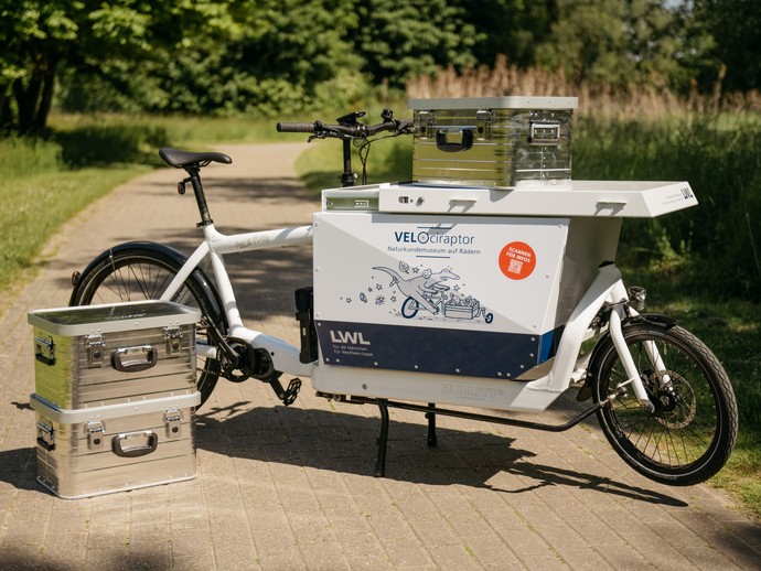 Ein E-Lastenrad mit der Aufschrift VELOciraptor und silberne Koffer auf einem Radweg im Grünen. (vergrößerte Bildansicht wird geöffnet)