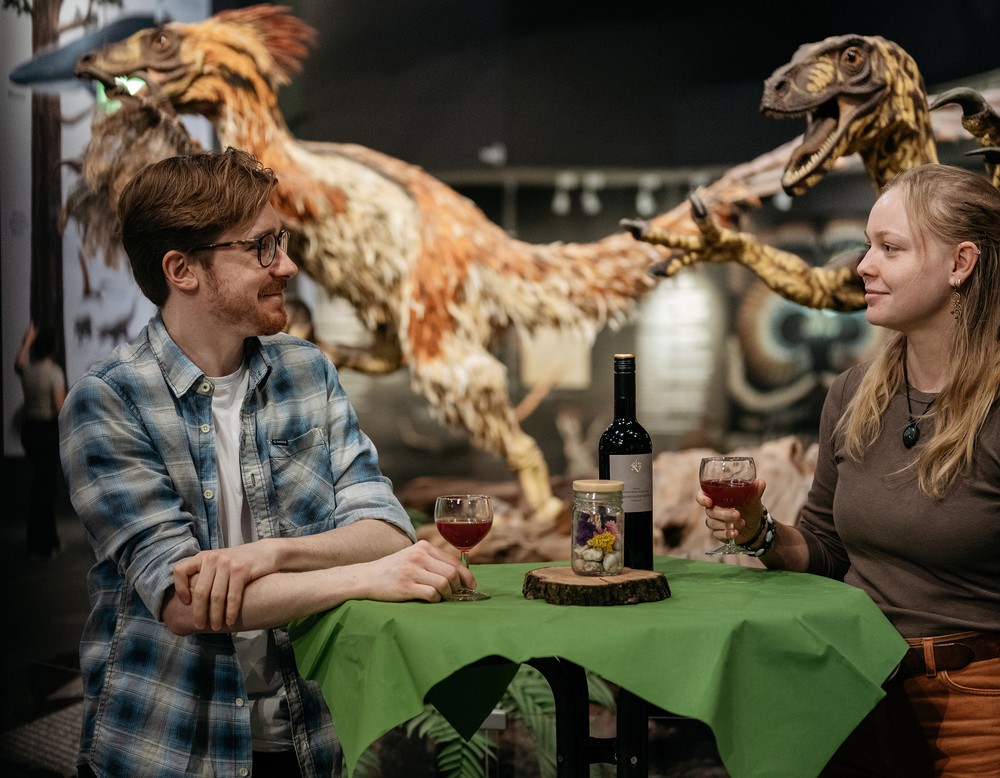 Ein Mann und eine Frau haben Weingläser in der Hand. Im Hintergrund stehen Dinosaurier-Modelle.